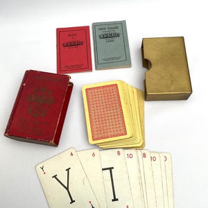 Könnte beinhalten: Ein Vintage-Lexikon-Kartenspiel-Set. Das Set enthält ein rotes Regelbuch, ein grünes Buch mit neuen Spielen, einen goldfarbenen Kartenhalter aus Metall, ein Kartenspiel mit rotem Muster auf der Rückseite sowie Buchstaben- und Zahlenkarten.