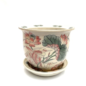May include: Cream ceramic plant pot with a matching saucer. The pot features a hand-painted design of pink lotus flowers, green leaves, and a red bird. The rim has small green leaf accents.