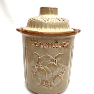 May include: Tan ceramic Rumtopf pot with a lid. The pot features embossed fruit and the word "Rumtopf" in script. The lid has a decorative pattern. The pot is designed for preserving fruit in rum.