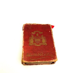 May include: A vintage, rectangular book with a distressed red cover. The cover features gold lettering and a crest with squirrels and the words "Confido" and "Gold Berry." A red ribbon marks a page. The book's title is "John Walker & Sons."