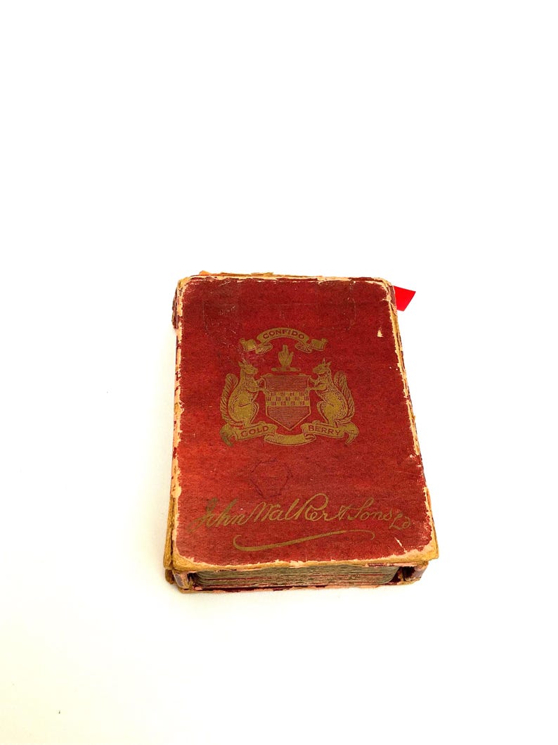 May include: A vintage, rectangular book with a distressed red cover. The cover features gold lettering and a crest with squirrels and the words "Confido" and "Gold Berry." A red ribbon marks a page. The book's title is "John Walker & Sons."