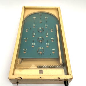 Könnte beinhalten: Vintage Flipper-Spiel aus Holz mit einem türkisfarbenen Spielfeld und nummerierten Bewertungsbereichen. Das Spiel hat eine Glasabdeckung, Metallkugeln und einen Ausziehhebel. Der Rahmen ist hellcremefarben.