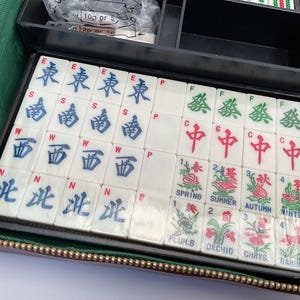 May include: A vintage Mahjong set with white tiles featuring Chinese characters and floral illustrations. The set includes tiles marked with East, South, West, and North, along with Spring, Summer, Autumn, and Winter symbols. The tiles are stored in a black case.