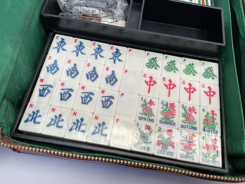 May include: A vintage Mahjong set with white tiles featuring Chinese characters and floral illustrations. The set includes tiles marked with East, South, West, and North, along with Spring, Summer, Autumn, and Winter symbols. The tiles are stored in a black case.