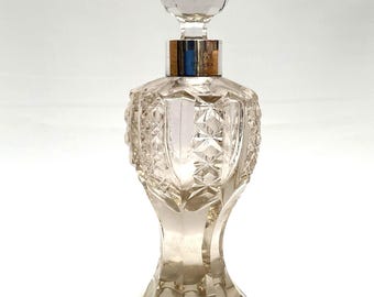 Può includere: Un flacone di profumo vintage trasparente con un tappo di cristallo sfaccettato. Il flacone ha un colletto color argento e un corpo in vetro strutturato e dettagliato. La base del flacone ha un design svasato decorativo. Il flacone è vuoto.