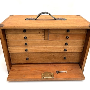 May include: Wooden toolbox with multiple drawers and a hinged front panel. The box has a leather handle and metal corner reinforcements. A small key rests on the open front panel, suggesting a locking mechanism. The drawers have black knobs.