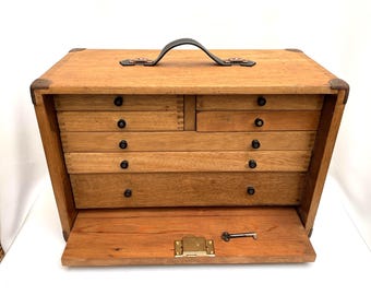 Könnte beinhalten: Holzwerkzeugkasten mit mehreren Schubladen und einer aufklappbaren Frontplatte. Die Box hat einen Ledergriff und Metallecken. Ein kleiner Schlüssel liegt auf der geöffneten Frontplatte. Die Schubladen haben schwarze Knöpfe.