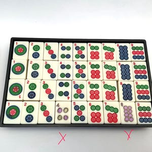 May include: A set of ivory Mahjong tiles in a black tray, displaying coloured symbols and numbers. A brown leather case with a gold zip is on the left. The game set is ready for play. The tiles are approximately 2.5 cm square.