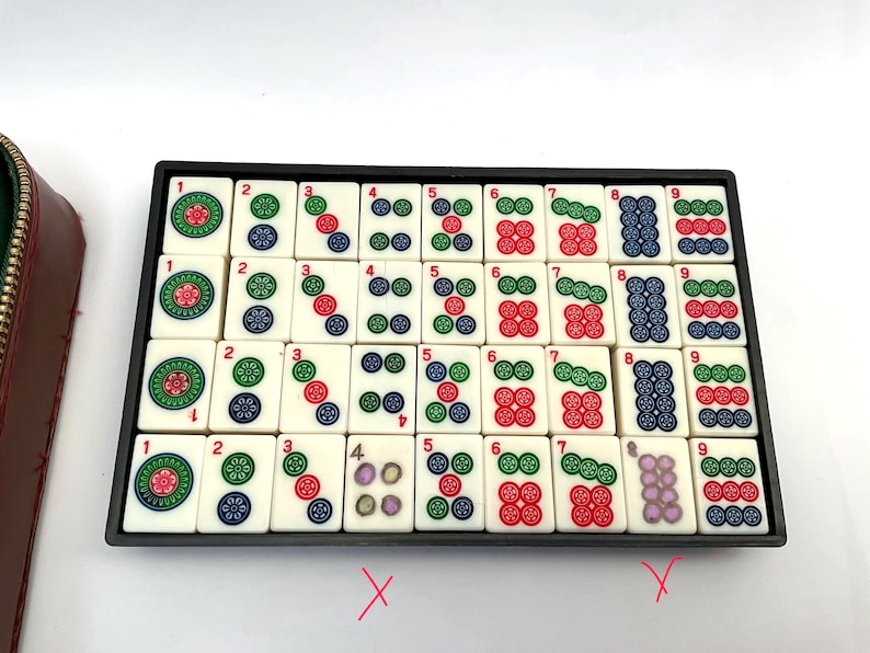 May include: A set of ivory Mahjong tiles in a black tray, displaying coloured symbols and numbers. A brown leather case with a gold zip is on the left. The game set is ready for play. The tiles are approximately 2.5 cm square.