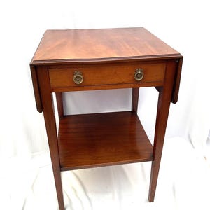 Puede incluir: Una pequeña mesa auxiliar de madera antigua con un cajón y un estante inferior. La mesa es de madera oscura con un acabado pulido. El cajón tiene dos pomos de latón. La mesa mide aproximadamente 61 cm de altura.