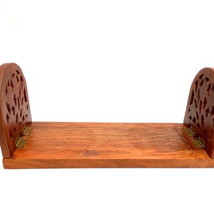 May include: Wooden book holder with a natural finish. Features two arched sides with carved leaf designs and a flat base. The sides are connected to the base with brass hinges, allowing it to fold flat.
