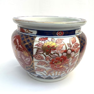 Puede incluir: Una maceta de cerámica redonda con base blanca decorada con complejos patrones florales en rojo, oro y azul. El diseño incluye peonías y follaje detallados, acentuados por un borde de rejilla geométrica azul oscuro y un borde dorado.