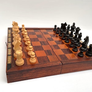 Puede incluir: Un juego de ajedrez de madera con un tablero plegable. El tablero es marrón con un patrón de cuadros. Las piezas de ajedrez son negras y blancas. El juego se muestra sobre una superficie blanca.