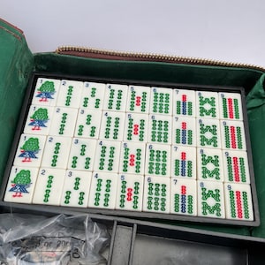 May include: A Mahjong set with white tiles displaying green, red, and blue symbols and numbers, arranged in a black tray. The tray is within a green and brown zippered case, indicating a game of strategy and skill.