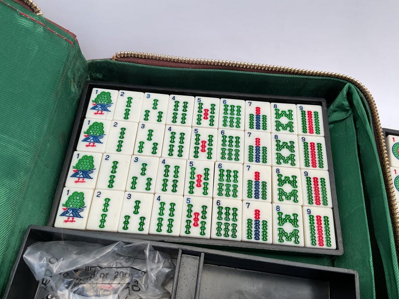 May include: A Mahjong set with white tiles displaying green, red, and blue symbols and numbers, arranged in a black tray. The tray is within a green and brown zippered case, indicating a game of strategy and skill.