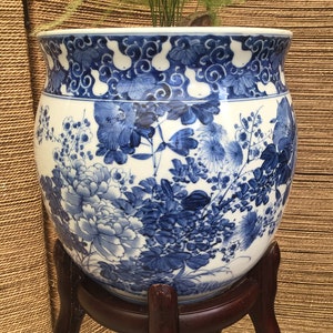 May include: A large, round, blue and white porcelain planter with a floral design. The planter is sitting on a dark wood stand with three legs.