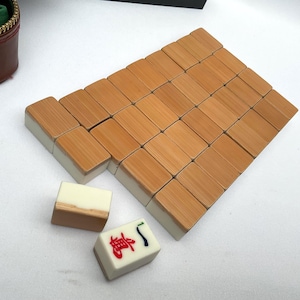 May include: A set of wooden Mahjong tiles with a light brown finish and white edges. The tiles are arranged in a rectangular pattern, with a few individual tiles nearby. A brown zippered case is visible in the background.