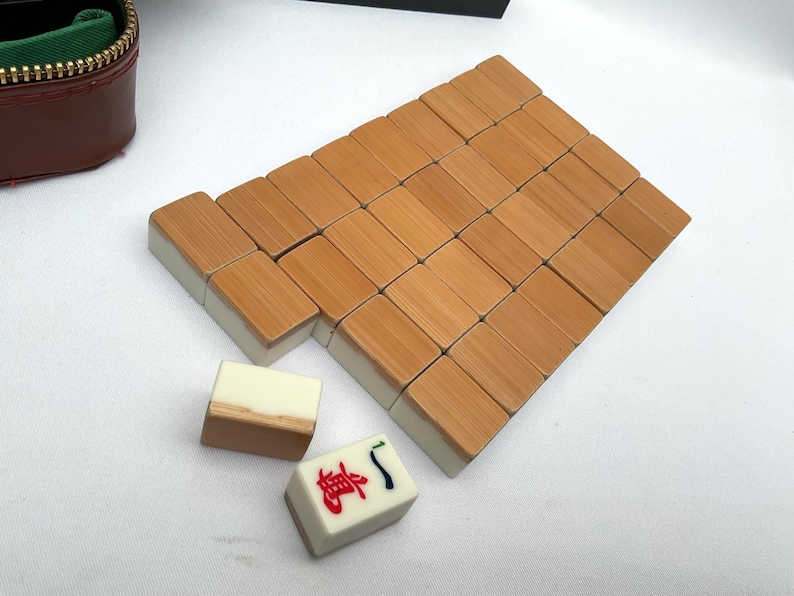 May include: A set of wooden Mahjong tiles with a light brown finish and white edges. The tiles are arranged in a rectangular pattern, with a few individual tiles nearby. A brown zippered case is visible in the background.