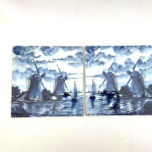 May include: Two square ceramic tiles featuring a blue and white windmill scene. The tiles show windmills, boats, and water under a cloudy sky. The artwork is reminiscent of Delftware pottery.