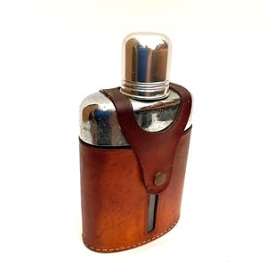 May include: A stainless steel hip flask with a polished metal cap, housed in a brown leather case. The leather holder features a button clasp and a small viewing window. The flask is designed for carrying liquids.