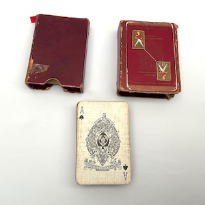 May include: An antique playing card with an Ace of Spades design, alongside two red playing card boxes. The card features a detailed black and white illustration. One box is open, and the other is closed, both showing signs of age.