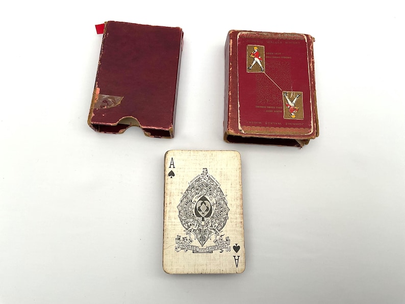 May include: An antique playing card with an Ace of Spades design, alongside two red playing card boxes. The card features a detailed black and white illustration. One box is open, and the other is closed, both showing signs of age.