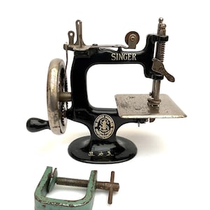 Könnte beinhalten: Eine schwarze Vintage-Singer-Handnähmaschine mit silberfarbenen Metallkomponenten, einer Nadelstange und einer flachen Platte. Die Maschine trägt das Singer-Logo und den Schriftzug. Eine separate grüne Metallschraubzwinge liegt darunter.