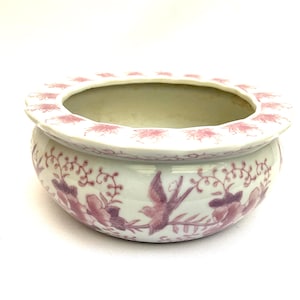 Op de afbeelding: Een witte keramische kom met een roze bloemen- en vogelontwerp. De rand heeft een herhalend roze bloemenpatroon. De kom is rond en heeft een brede opening, geschikt voor decoratief of functioneel gebruik.