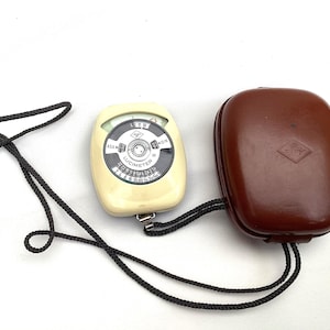 Könnte beinhalten: Ein Vintage-Belichtungsmesser mit cremefarbenem Gehäuse und schwarzem Zifferblatt, verbunden mit einem braunen Lederetui durch eine schwarze Kordel. Der Belichtungsmesser hat ein rundes Zifferblatt mit Zahlen und Markierungen. Das Etui hat ein rautenförmiges Logo.