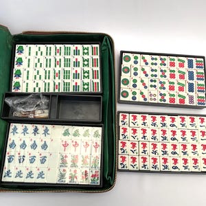 May include: A Mahjong set with ivory tiles, displaying green, red, and blue symbols and characters. The set includes a green zippered case, dice, and tile racks. The tiles are arranged in black trays.