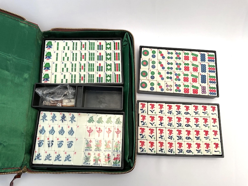 May include: A Mahjong set with ivory tiles, displaying green, red, and blue symbols and characters. The set includes a green zippered case, dice, and tile racks. The tiles are arranged in black trays.