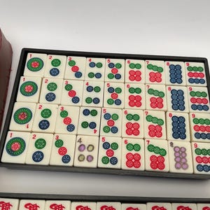 May include: A Mahjong set featuring ivory tiles with colourful symbols and numbers, arranged in a black tray. A brown leatherette case is visible. The tiles display red, green, blue, and purple markings. The game set is designed for strategic tile-matching.