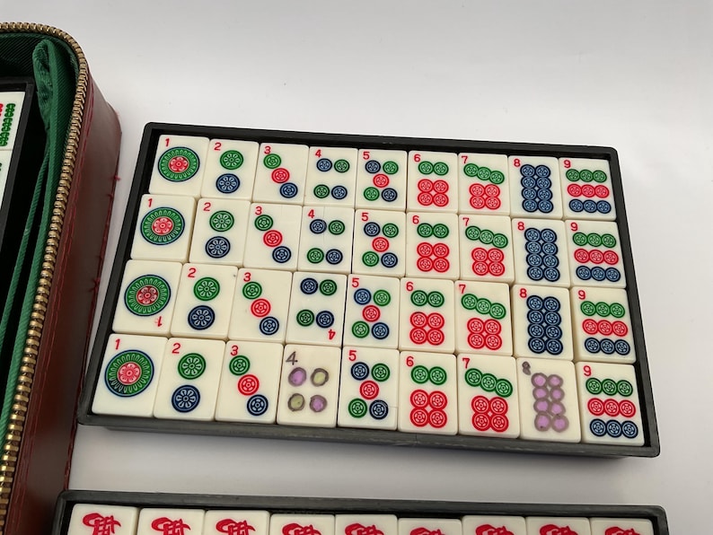May include: A Mahjong set featuring ivory tiles with colourful symbols and numbers, arranged in a black tray. A brown leatherette case is visible. The tiles display red, green, blue, and purple markings. The game set is designed for strategic tile-matching.