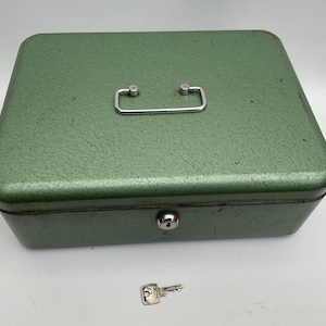 May include: A vintage green metal cash box with a silver handle and a key lock. The box has a textured surface and a small key is placed in front of it. The cash box is rectangular and has rounded corners.