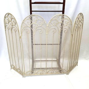 May include: A cream-coloured, three-panel folding fireplace screen made of metal mesh. Each panel features a rounded top with decorative scrollwork and vertical bars. The screen is hinged to allow for adjustable positioning in front of a fireplace.
