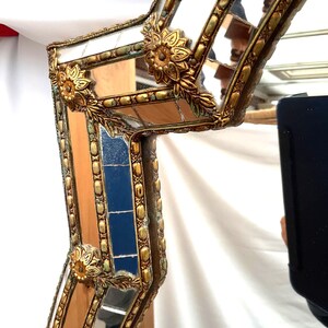 K&ouml;nnte beinhalten: Nahaufnahme eines dekorativen Spiegelrahmens mit Goldfinish. Der Rahmen weist verzierte florale Akzente und ein geometrisches Design mit verspiegelten Abschnitten auf. Der Rahmen hat eine Vintage-&Auml;sthetik und ist vor einem wei&szlig;en Hintergrund platziert.