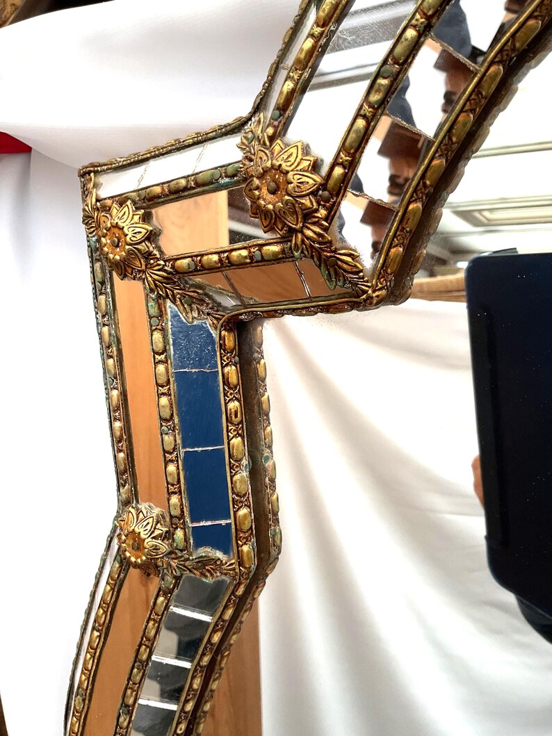 K&ouml;nnte beinhalten: Nahaufnahme eines dekorativen Spiegelrahmens mit Goldfinish. Der Rahmen weist verzierte florale Akzente und ein geometrisches Design mit verspiegelten Abschnitten auf. Der Rahmen hat eine Vintage-&Auml;sthetik und ist vor einem wei&szlig;en Hintergrund platziert.