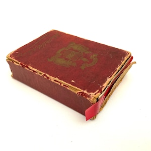 May include: A small, antique book with a distressed red cover and gold lettering. The book has a decorative emblem on the cover and a red ribbon bookmark. The edges of the book show signs of wear, suggesting its age.