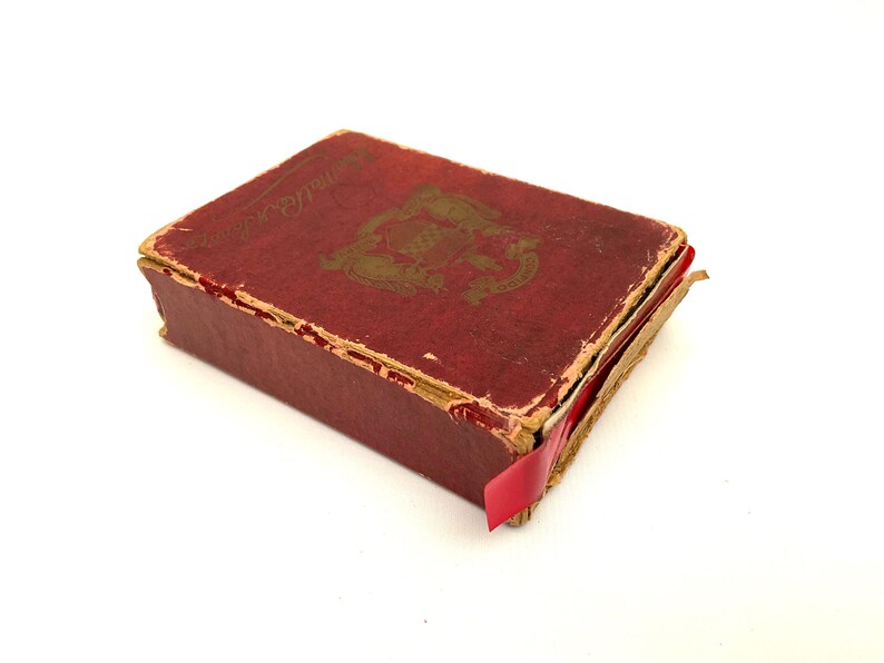 May include: A small, antique book with a distressed red cover and gold lettering. The book has a decorative emblem on the cover and a red ribbon bookmark. The edges of the book show signs of wear, suggesting its age.