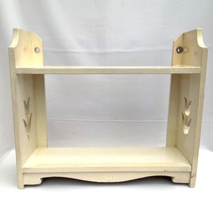 Puede incluir: Estantería de madera crema de dos niveles con recortes decorativos y base curva. La estantería tiene dos niveles para exhibir objetos y está diseñada con una estética clásica y vintage. La estantería mide aproximadamente 61 cm de ancho.