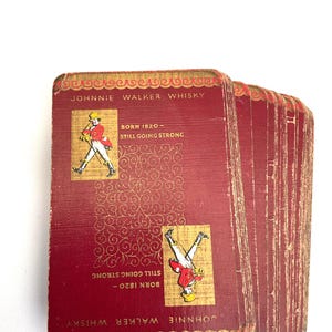 May include: A deck of vintage Johnnie Walker Whisky playing cards. The cards are a deep red with gold accents and feature the Johnnie Walker logo and text. The cards read "Born 1820 - Still Going Strong."