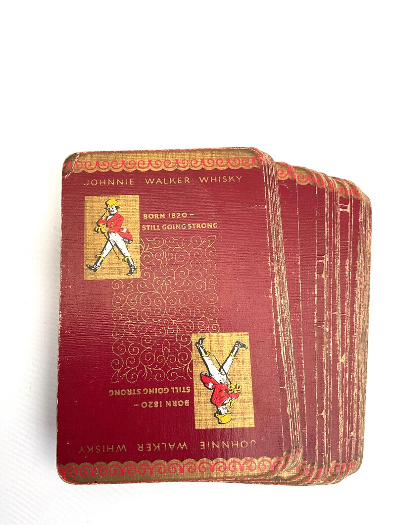 May include: A deck of vintage Johnnie Walker Whisky playing cards. The cards are a deep red with gold accents and feature the Johnnie Walker logo and text. The cards read "Born 1820 - Still Going Strong."