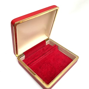 May include: Open red jewelry box with a gold trim. The interior is lined with red velvet and a cream-colored lid. The box is square and has a hinged lid. The box is empty.