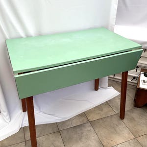 May include: A mint green drop-leaf table with a rectangular top and four brown wooden legs. The table is shown with the leaf down. The table's dimensions are approximately 76 cm long, 46 cm wide, and 71 cm high.