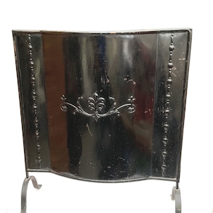 May include: A black fireplace screen with a decorative floral design in the center. The screen has a curved top and is supported by two curved legs. The screen has vertical beaded accents on each side.