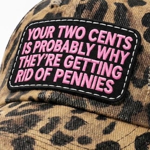 May include: A leopard print baseball cap with a black mesh back and a rectangular patch on the front. The patch features the pink embroidered text: YOUR TWO CENTS IS PROBABLY WHY THEY'RE GETTING RID OF PENNIES. The cap has a distressed brim.