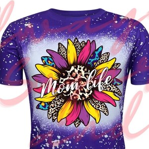 Puede incluir: Camiseta morada con un diseño de pintura salpicada blanca. Un girasol colorido con detalles de estampado de leopardo se encuentra en el centro de la camiseta. El texto "Mom life" está dentro del girasol.