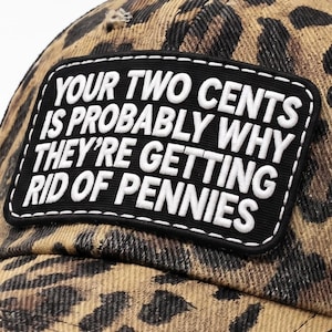 May include: A leopard print baseball cap with a black mesh back and a rectangular black patch on the front. The patch features white text that reads: YOUR TWO CENTS IS PROBABLY WHY THEY'RE GETTING RID OF PENNIES. The cap has a distressed brim.
