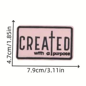 May include: A rectangular patch with the words "CREATED with a purpose" in black lettering on a light pink background. The patch measures 3.11 inches wide and 1.85 inches high.