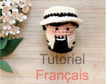 May include: A crocheted amigurumi doll wearing a white hat with black stripes. The doll has a black beard and a pink face with black eyes and a white smile. The doll is sitting on a wooden surface with the text "Tutoriel Français" below it.